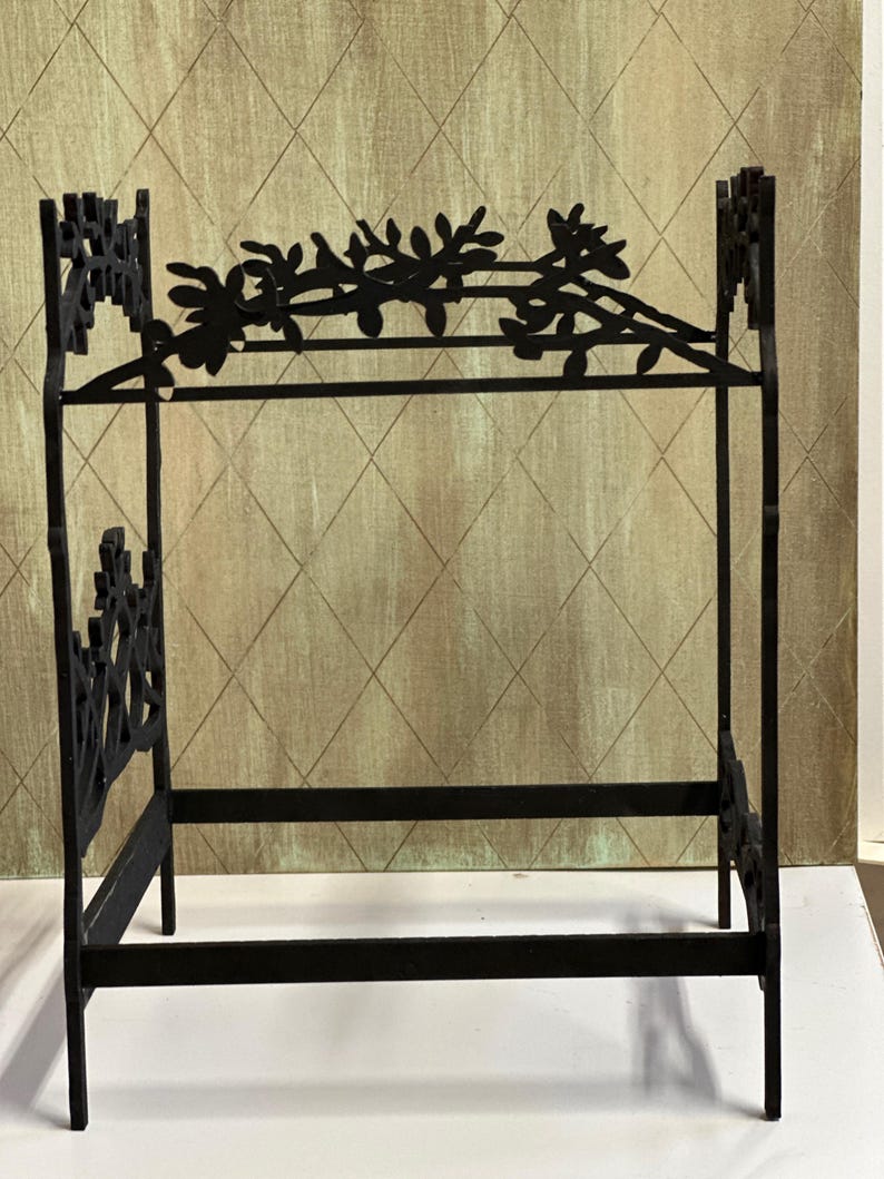 May include: A black metal doll bed with a floral design on the headboard and footboard. The bed frame is rectangular with a horizontal bar for the mattress. The headboard features a decorative floral vine.