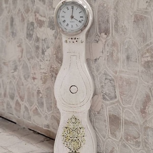 Puede incluir: Un reloj de madera blanco con una esfera blanca y números romanos negros. El reloj tiene la forma de un jarrón alto y delgado con un diseño floral decorativo.