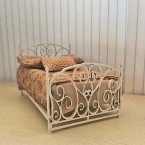 May include: White metal bed frame with a decorative scroll design. The bed is made for a dollhouse or miniature setting.