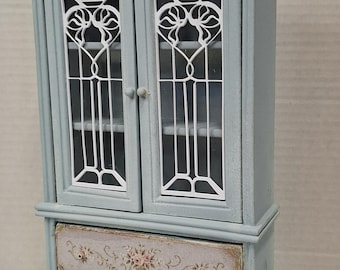 May include: A light blue miniature cabinet featuring two glass doors with white decorative overlays and a single bottom drawer. The drawer front is decorated with a floral pattern in pink and green tones. The cabinet stands on four small ornate feet.