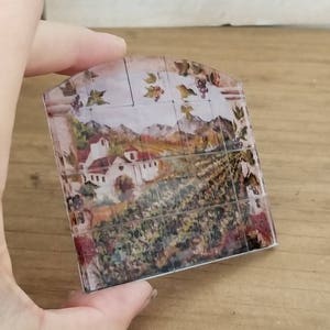 May include: A decorative tile featuring a vintage-style image of a vineyard and a small village in the background. The tile is made of glass and has a slightly curved shape.