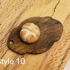 May include: A small, round, brown and white bread roll on a dark brown wooden cutting board with a scalloped edge. The cutting board has a hole in the top for hanging. The background is a light brown wooden surface. Style 10.