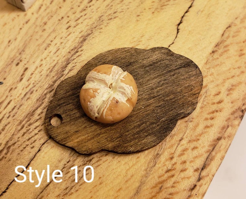 May include: A small, round, brown and white bread roll on a dark brown wooden cutting board with a scalloped edge. The cutting board has a hole in the top for hanging. The background is a light brown wooden surface. Style 10.