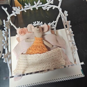 May include: A whimsical miniature bed scene featuring a fabric mouse doll wearing an orange dress, nestled in a bed with pink bedding and a cream-colored knitted blanket. The bed frame is white with a decorative branch design.