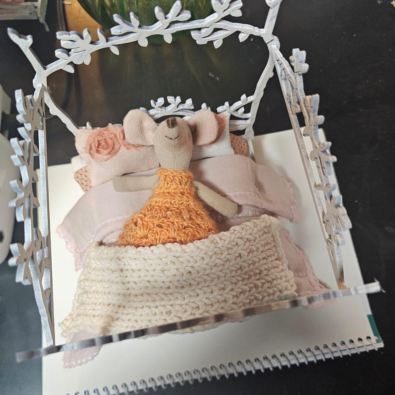 May include: A whimsical miniature bed scene featuring a fabric mouse doll wearing an orange dress, nestled in a bed with pink bedding and a cream-colored knitted blanket. The bed frame is white with a decorative branch design.
