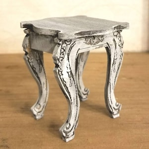May include: A small, white, wooden table with ornate legs and a distressed finish. The table has a square top and is perfect for a small space.