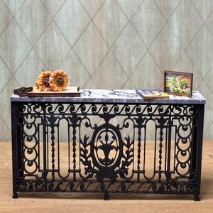 May include: A black wrought iron console table with a marble top. The table has a decorative design with a central oval and floral motifs. There are two small items on the table, a picture frame and a book.