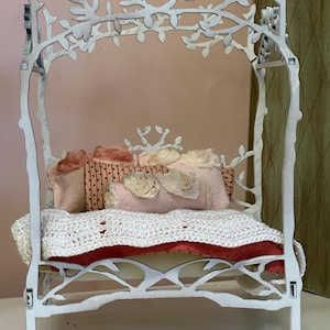May include: A white, decorative miniature bed with a branch-like frame. The bed is adorned with pink and cream-colored pillows, a white crocheted blanket, and a red cushion. The bed frame has a whimsical, nature-inspired design.