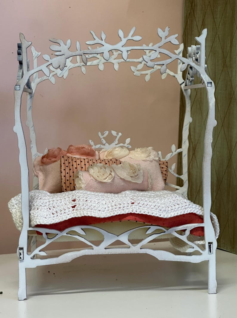May include: A white, decorative miniature bed with a branch-like frame. The bed is adorned with pink and cream-colored pillows, a white crocheted blanket, and a red cushion. The bed frame has a whimsical, nature-inspired design.