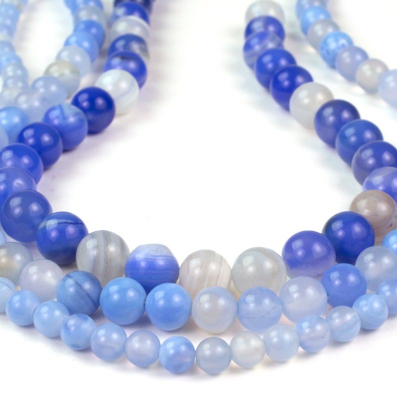 May include: A strand of blue agate beads in various shades of blue, from light to dark, with white striations. The beads are round and smooth, and they are strung on a clear thread.