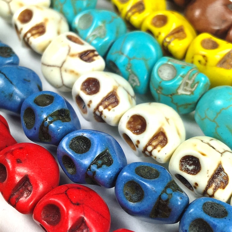 Puede incluir: Una hebra de cuentas de calavera de colores en turquesa, blanco, rojo y azul. Las cuentas tienen todas la forma de una calavera y tienen una textura ligeramente rugosa.