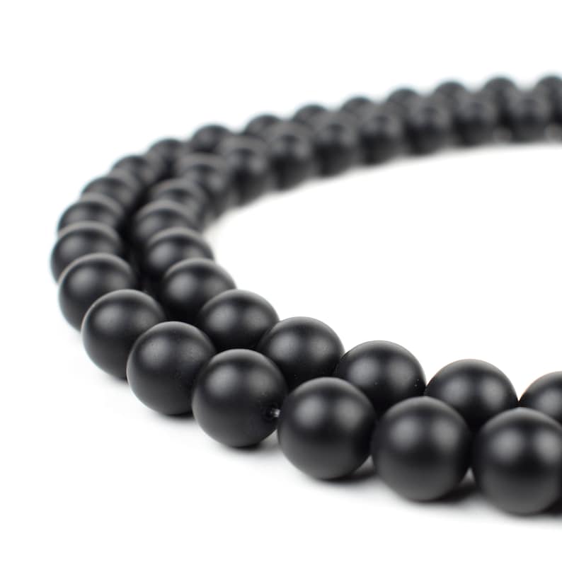 May include: A strand of black matte round beads, arranged in a circle on a white background.