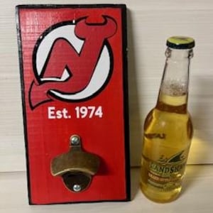 May include: A red wooden bottle opener with the text "Est. 1974" and a logo. Next to it is a clear glass bottle filled with a golden liquid. The bottle has a yellow cap and a label.