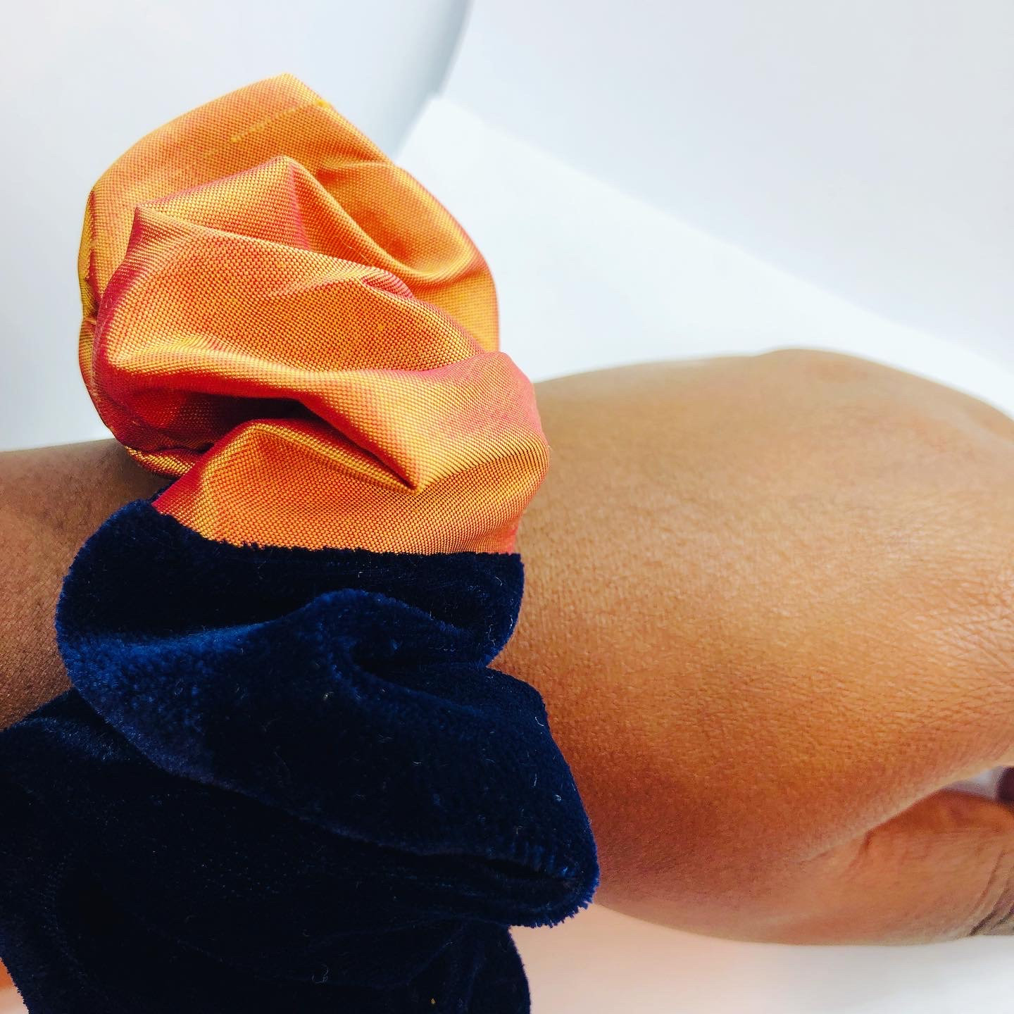 May include: A two-tone scrunchie in orange and navy blue. The top section is a bright orange fabric, and the bottom is a deep navy blue velvet. This hair accessory is designed to hold hair.