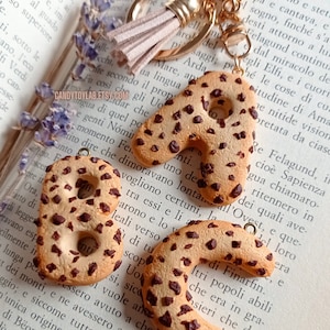 May include: A gold keychain with a brown tassel charm. The keychain features two large, brown cookie-shaped letters, "A" and "B", with chocolate chips. The letters are attached to the keychain with a gold ring.