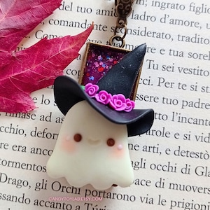 May include: A white ghost charm with a black witch hat and pink flowers. The charm is hanging from a gold chain with a purple and glitter filled pendant.