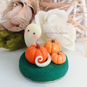 Puede incluir: Una figurita de fantasma blanco con una cara amigable se sienta sobre una base verde con tres calabazas naranjas. Las calabazas están dispuestas alrededor del fantasma, con la calabaza más grande en el centro. La base está hecha de un material texturizado que se asemeja a la hierba. El fantasma tiene una pequeña cola rizada.