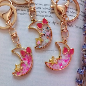 May include: Three gold crescent moon keychains with pink bows and glitter inside. Each keychain has a different color glitter and a gold star.