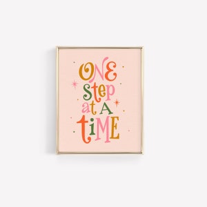 May include: A framed print with a pink background and gold frame. The print features the text "ONE step at a time" in colorful, stylized lettering.