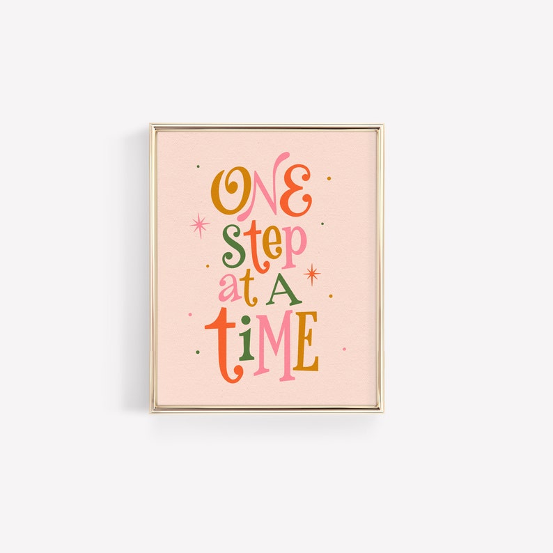 May include: A framed print with a pink background and gold frame. The print features the text "ONE step at a time" in colorful, stylized lettering.