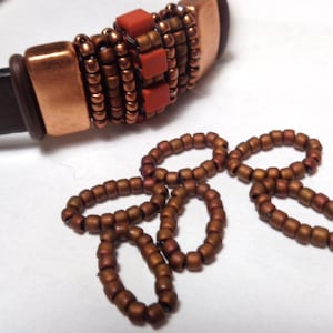 May include: A brown leather bracelet with copper-colored metal accents and small, reddish-brown beads. The bracelet features a decorative section with square, reddish-brown beads and a series of smaller beads.