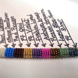 May include: A close-up of a beaded bracelet with different colored beads. The beads are labeled with names like "Pearl Lustered", "Opaque Consflower", "Red Picasso", "Pf Gold", "Pt Violet", "Fushia Lived", "Antique Brass", "Picasso Turquoise", "Lapis Blue Rosso", and "Pf Silver".