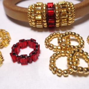 May include: An assortment of beaded jewelry, including rings and a bracelet. The rings are in gold and red, with various bead shapes. The bracelet features gold beads with a red center. The jewelry is displayed on a white surface.