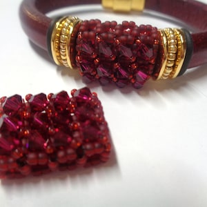 May include: A burgundy leather bracelet with a gold clasp and accents. The bracelet features a central section of clustered, faceted red beads. A matching rectangular bead component is also present.