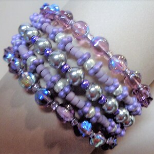 May include: A multi-strand bracelet with purple, lavender, and iridescent beads. The bracelet is made with a variety of beads, including small round beads, larger round beads, and faceted beads.