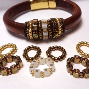May include: A brown leather bracelet with a gold magnetic clasp and a decorative band of gold, brown, and white beads. The bracelet is surrounded by eight smaller bead rings in various colors and patterns.
