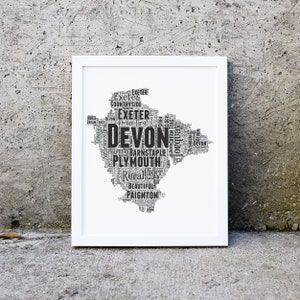 May include: A framed wall art print featuring a map of Devon, England, composed entirely of black text on a white background. The typography includes place names like Exeter, Plymouth, Torquay, Barnstaple, and Paignton, along with phrases like Cider.