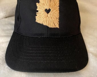 Op de afbeelding: Een zwarte baseballpet met een geborduurde omtrek van de staat Arizona in tan met een zwart hart in het midden.