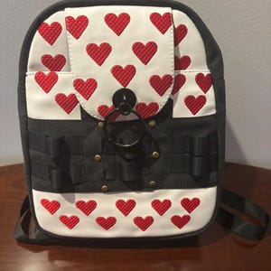 May include: A black and white backpack featuring a repeating pattern of embroidered red hearts. The front flap includes a large black circular metal ring closure, and the lower section has black nylon webbing straps secured with small brass-colored rivets.
