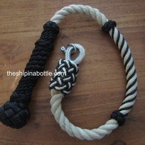 May include: A nautical rope accessory with a black and white design. The rope is made of thick, twisted fibers, with a black handle and a loop featuring a metal clasp. The text "theshipinabottle.com" is visible.