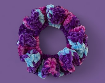 May include: A plush scrunchie in shades of purple, pink, and blue. The hair accessory is made of a soft, velvety material and has a ruffled design. The scrunchie is circular with a hole in the center, perfect for securing hair.
