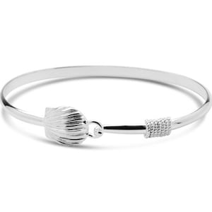 May include: A silver-colored bangle bracelet with a seashell-shaped clasp. The bracelet has a hook closure and a decorative, textured section near the clasp. The jewelry is displayed on a white background.