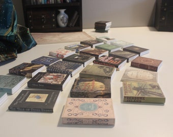 May include: A collection of miniature books with various titles, including "Oliver Twist", "The Tale of Peter Rabbit", "Treasure Island", and "Red-Fox". The books are arranged in a triangular shape on a white surface.
