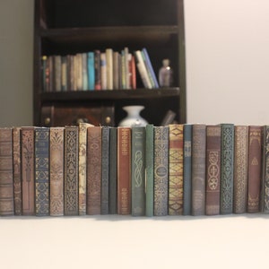 Puede incluir: Una fila de libros de tapa dura de estilo vintage, de color marrón y verde con lomos decorativos. Los libros están dispuestos en línea sobre una superficie blanca.