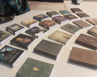 May include: A collection of miniature books with various titles and designs. The books are arranged on a white surface and feature different colors and patterns, including brown, green, and gold.