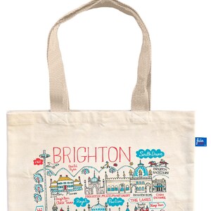 May include: Cream-colored tote bag with a colorful illustration of Brighton landmarks. The design includes the text "BRIGHTON" in red, along with drawings of buildings, the Brighton Racecourse, and the text "HOVE". The bag has beige handles.