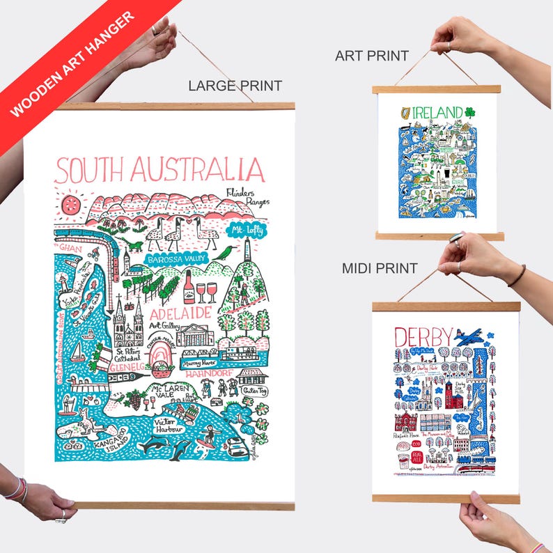 May include: Three wooden art hangers displaying illustrated maps of South Australia, Ireland, and Derby. Each print features colourful hand-drawn landmarks, text labels, and geographical details. The hangers consist of two light wood bars with a string.