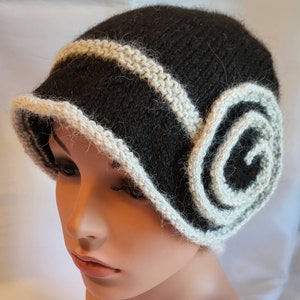 May include: A black and white knitted hat with a large white swirl on the side. The hat has a wide brim and is made of a soft, fuzzy yarn.