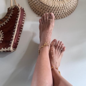 May include: Gold-colored foot jewelry with delicate chain and lace-like designs. The jewelry is worn on the feet and ankles. A brown woven bag hangs in the background.