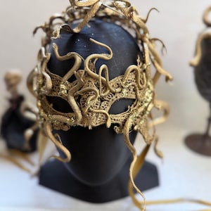 May include: A gold-colored lace masquerade mask featuring multiple snake-shaped embellishments extending from the frame. The mask is displayed on a black mannequin head, showcasing intricate metallic textures and winding serpent forms.