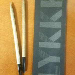 May include: Two wooden knitting needles with black tips. The needles are on a wooden surface next to a black card with the word "LYKKE" printed in white.