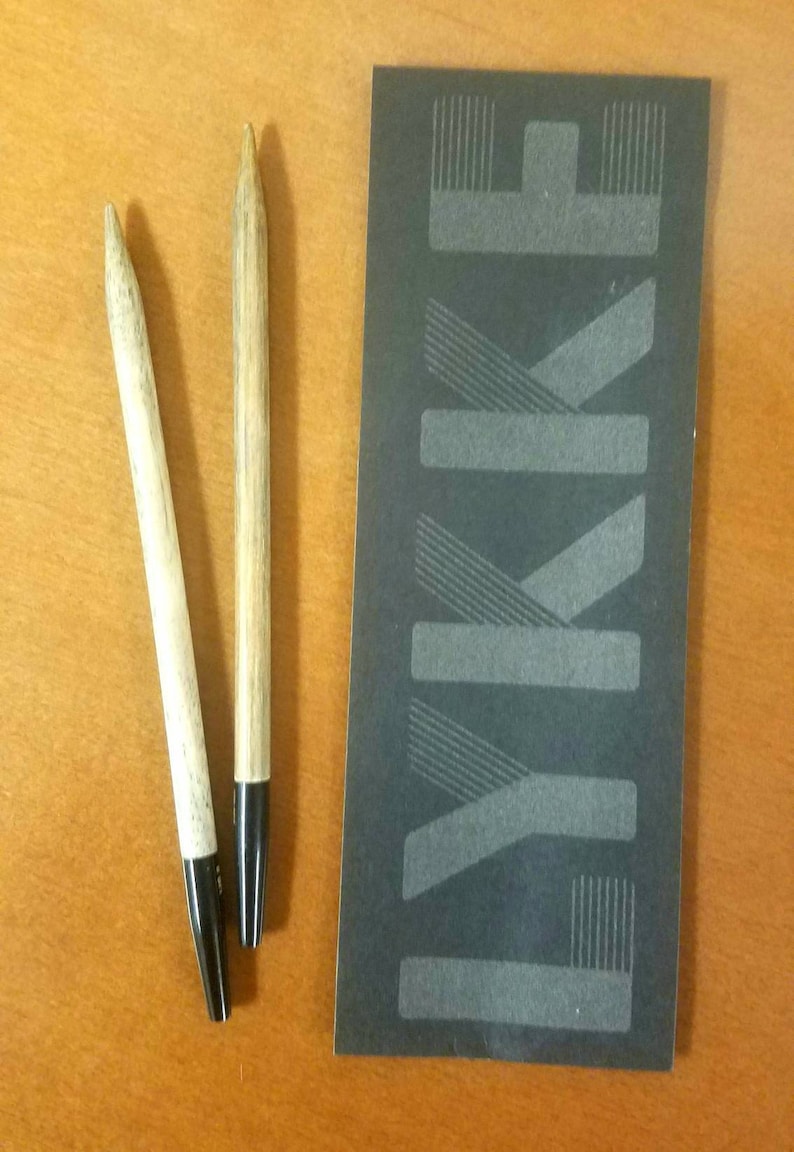 May include: Two wooden knitting needles with black tips. The needles are on a wooden surface next to a black card with the word "LYKKE" printed in white.