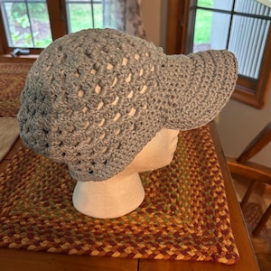 May include: A light blue crocheted hat with a visor, displayed on a white mannequin head. The hat has an open weave design and a rounded crown. The visor is solid and provides shade. The hat is likely handmade and suitable for sun protection.