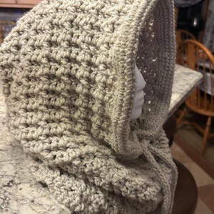 May include: A beige crocheted cowl with a hood. The cowl is made with a textured stitch and has a long, flowing tie.