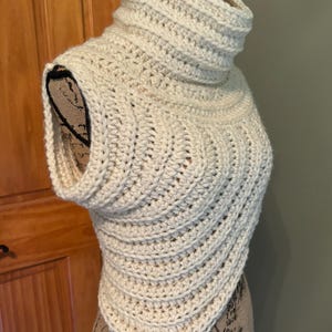 May include: A cream-colored crocheted cowl with a high neck and sleeveless design. The cowl is displayed on a mannequin.