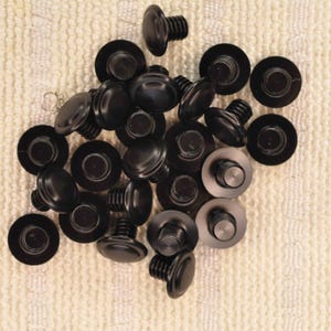 May include: A collection of black furniture screw buttons with a rounded head and threaded shaft. The buttons are scattered on a textured, off-white surface. The screw buttons are likely used for furniture assembly or repair.